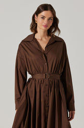 A woman is wearing a long-sleeved, brown shirt dress with a pointed collar. The dress features a button-down front and is cinched at the waist with a matching brown belt that has a metallic D-ring buckle. The skirt of the dress is gathered and pleated, creating a voluminous silhouette.